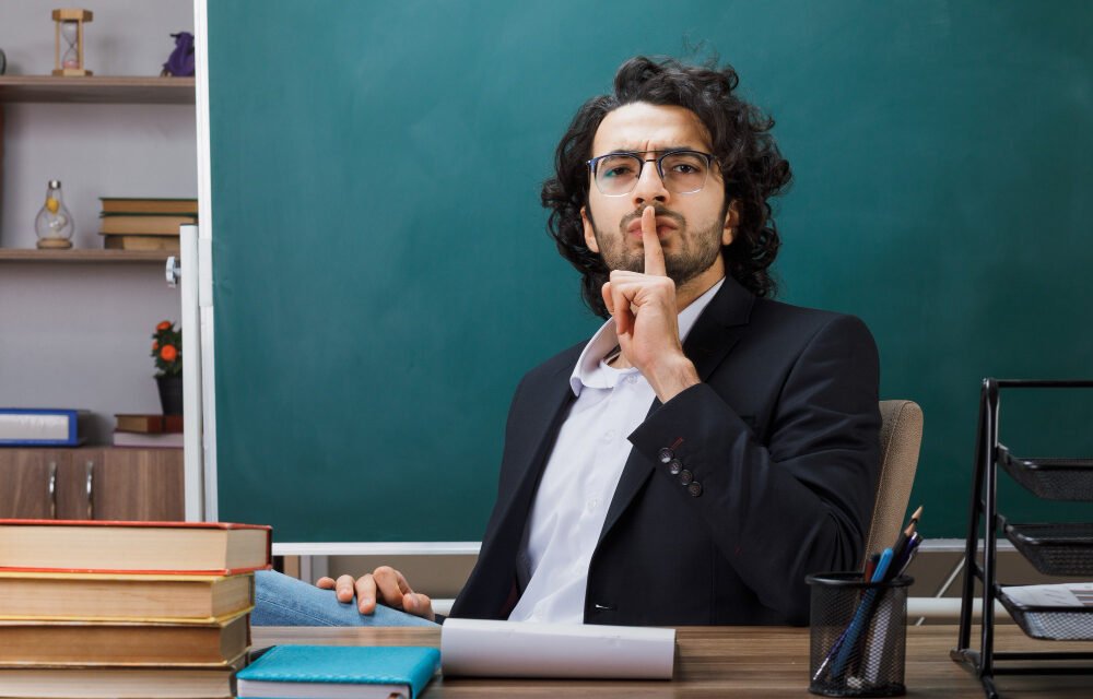 Male teacher in classroom gesturing for silence with finger on lips, surrounded by books and chalkboard, symbolizing discipline and focus in education