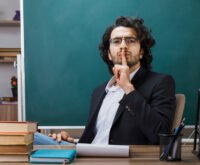 Male teacher in classroom gesturing for silence with finger on lips, surrounded by books and chalkboard, symbolizing discipline and focus in education