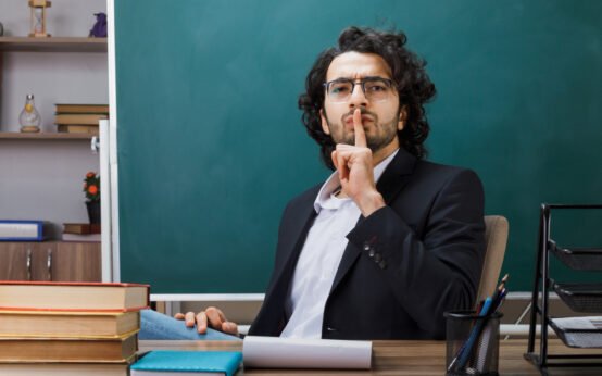 Male teacher in classroom gesturing for silence with finger on lips, surrounded by books and chalkboard, symbolizing discipline and focus in education
