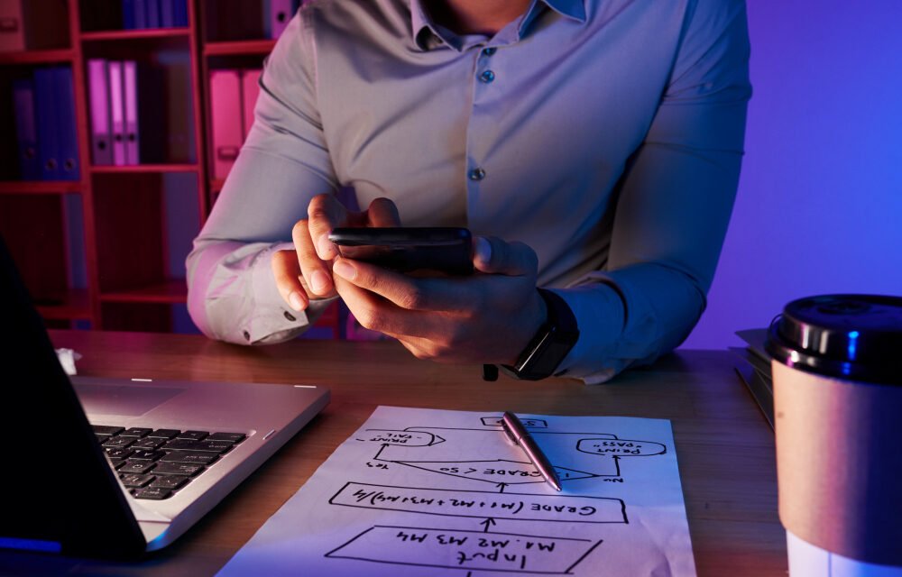 Entrepreneur reviewing business strategy on smartphone with laptop and startup planning notes on desk