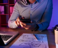Entrepreneur reviewing business strategy on smartphone with laptop and startup planning notes on desk