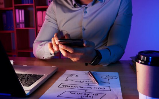 Entrepreneur reviewing business strategy on smartphone with laptop and startup planning notes on desk