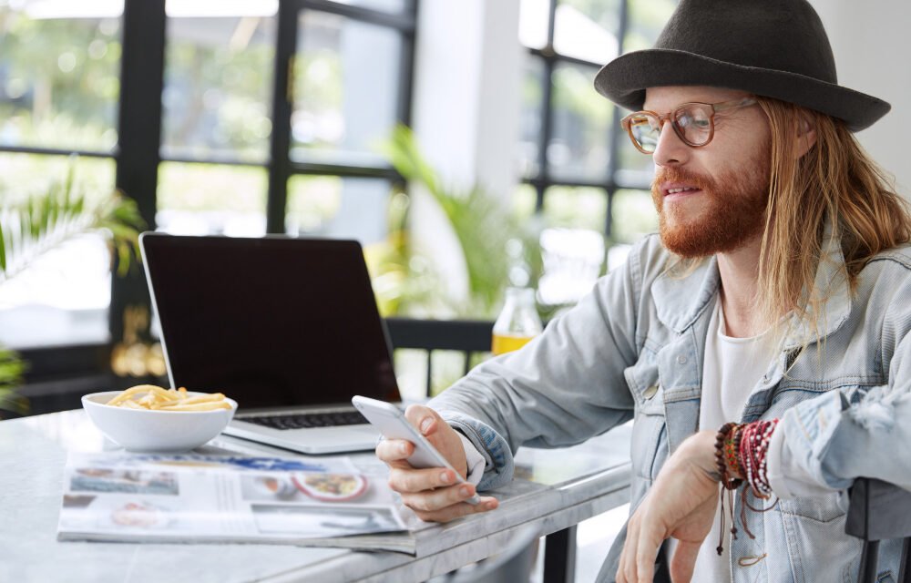 Trendy young man with long hair and hat using smartphone at cafe table with laptop, magazine, and snack bowl