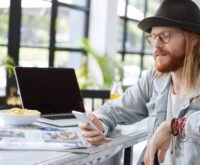 Trendy young man with long hair and hat using smartphone at cafe table with laptop, magazine, and snack bowl
