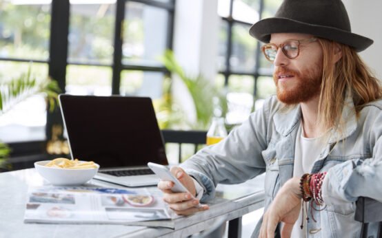 Trendy young man with long hair and hat using smartphone at cafe table with laptop, magazine, and snack bowl