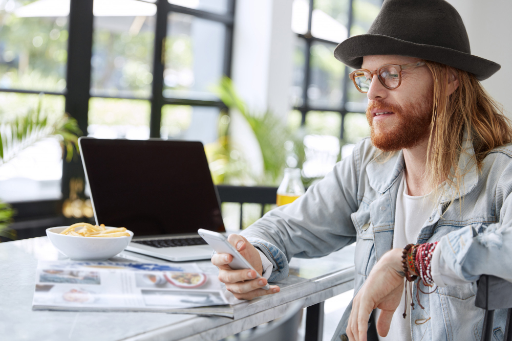 Trendy young man with long hair and hat using smartphone at cafe table with laptop, magazine, and snack bowl