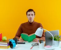 Focused student reading a book at a colorful study desk with educational supplies, laptop, and yellow background for e-learning or academic concept