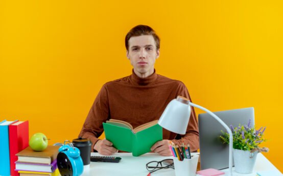 Focused student reading a book at a colorful study desk with educational supplies, laptop, and yellow background for e-learning or academic concept