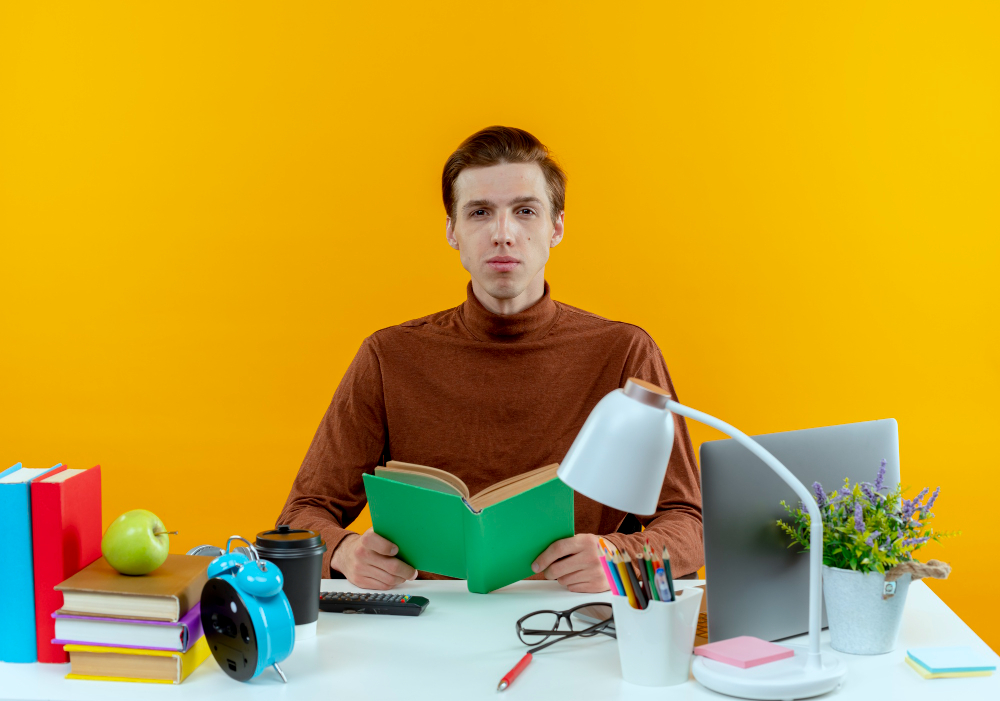 Focused student reading a book at a colorful study desk with educational supplies, laptop, and yellow background for e-learning or academic concept