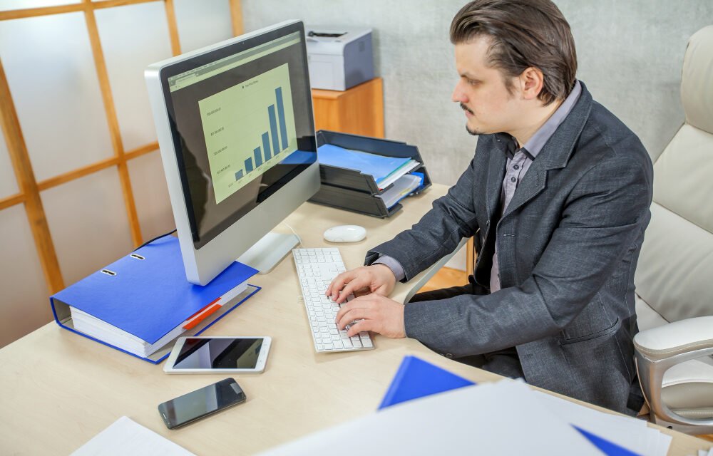 Businessman analyzing bar chart data on a desktop computer in a modern office setting, with documents, smartphone, and tablet on the desk