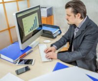 Businessman analyzing bar chart data on a desktop computer in a modern office setting, with documents, smartphone, and tablet on the desk