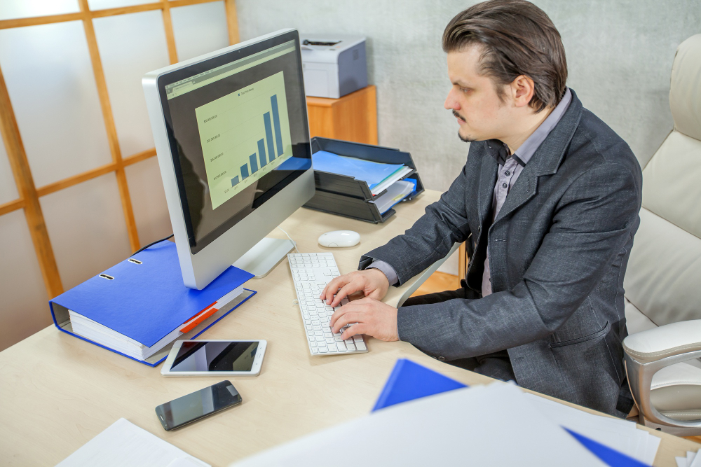 Businessman analyzing bar chart data on a desktop computer in a modern office setting, with documents, smartphone, and tablet on the desk