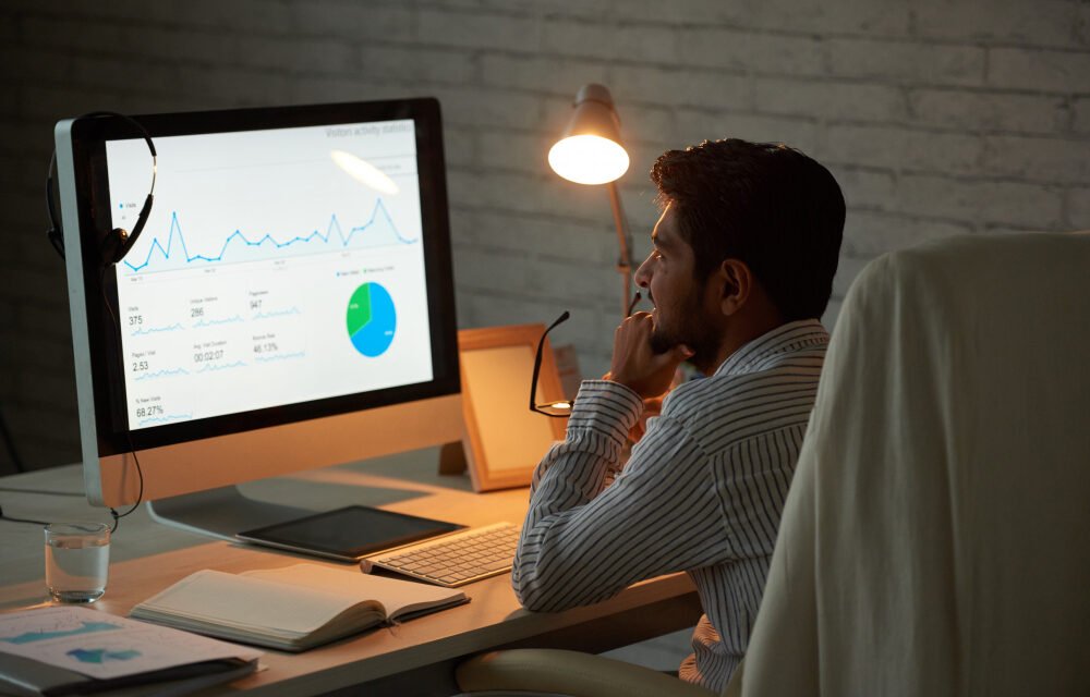 Business analyst reviewing financial performance graphs and pie chart on desktop monitor in low-light office environment