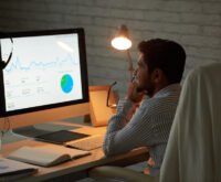 Business analyst reviewing financial performance graphs and pie chart on desktop monitor in low-light office environment