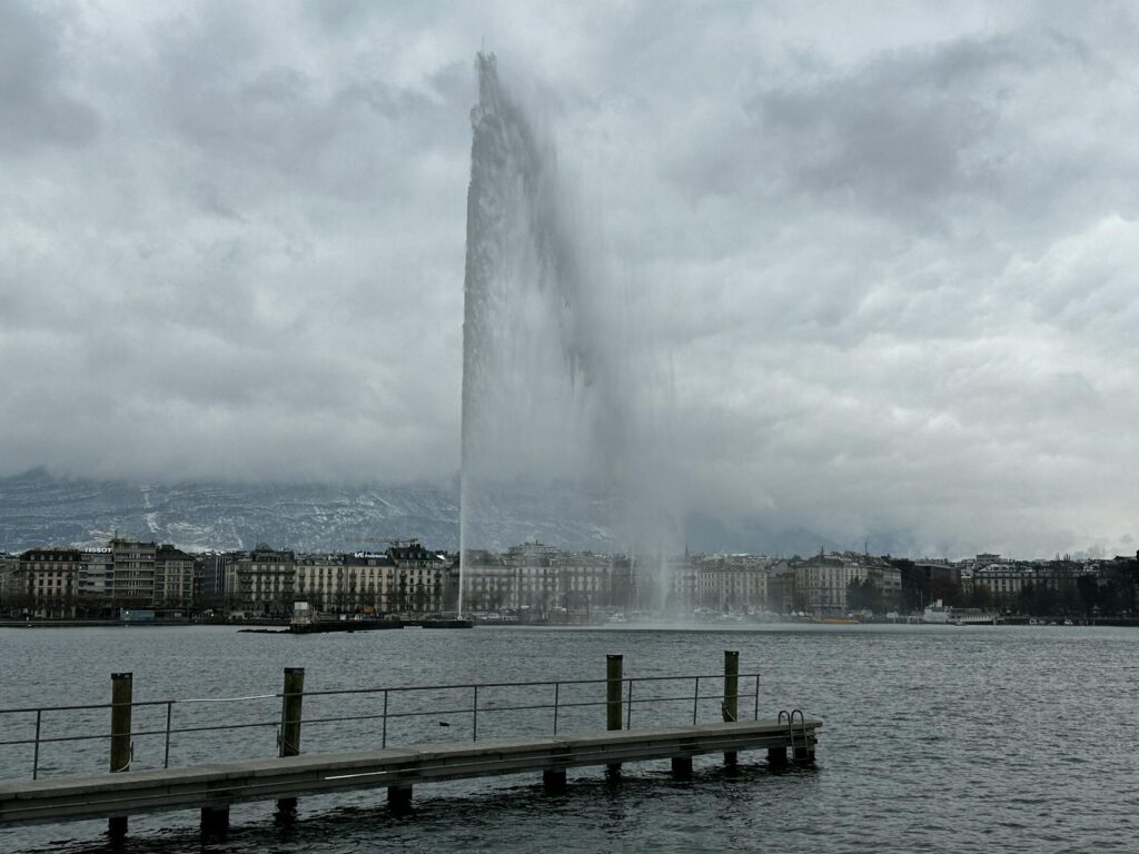 The iconic Jet d'Eau fountain spraying high into the air over Lake Geneva, with the city and mountains in the background.