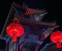 A traditional wooden boat drifts down a Suzhou canal illuminated by glowing red lanterns at dusk.