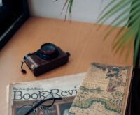 A person's hands writing on a blank postcard next to a vintage film camera and a cup of coffee.