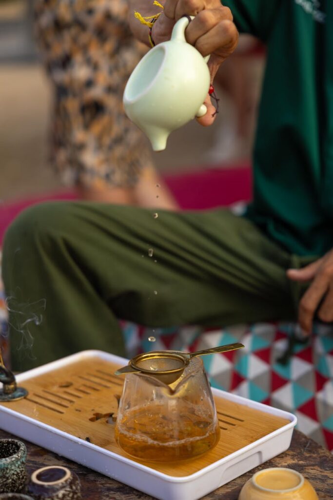 A person's hands carefully pouring steaming water from a kettle into a clear glass teapot, where vibrant red hibiscus tea is beginning to steep.