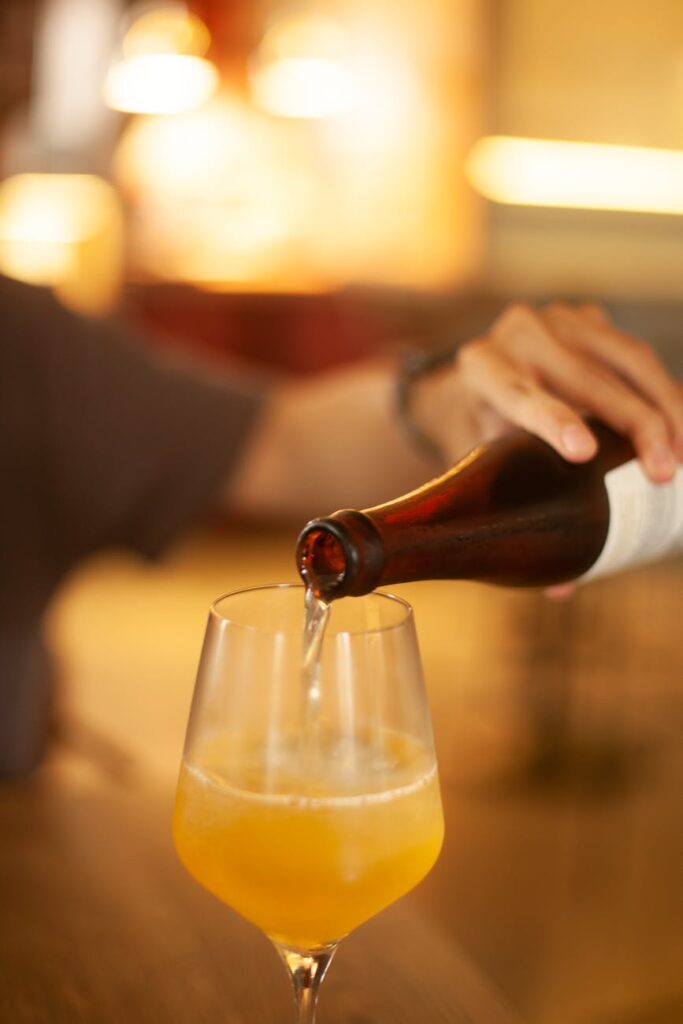 Amber-colored kombucha being poured from a bottle into a clear glass, with fizz and bubbles visible.
