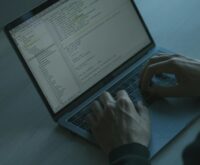 A hooded figure representing a hacker types code on a laptop, illustrating DeFi security risks.