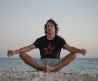 A large, diverse group of people practicing a standing yoga pose on a sandy beach during a vibrant sunset, all wearing immersive silent disco headphones.