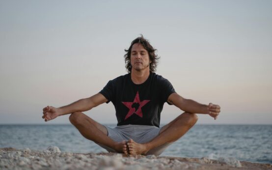 A large, diverse group of people practicing a standing yoga pose on a sandy beach during a vibrant sunset, all wearing immersive silent disco headphones.
