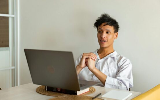 A young professional smiling while working on a laptop in a cozy, sunlit home office.