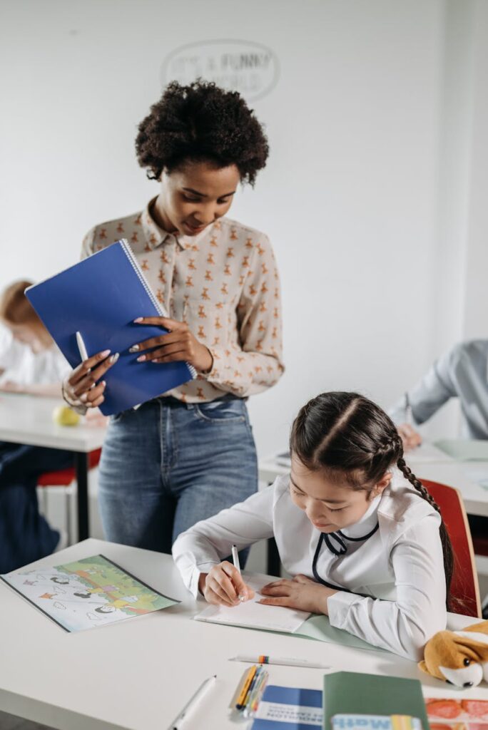A friendly female teacher points to a whiteboard while a small, helpful robot assistant stands beside her in a bright classroom.