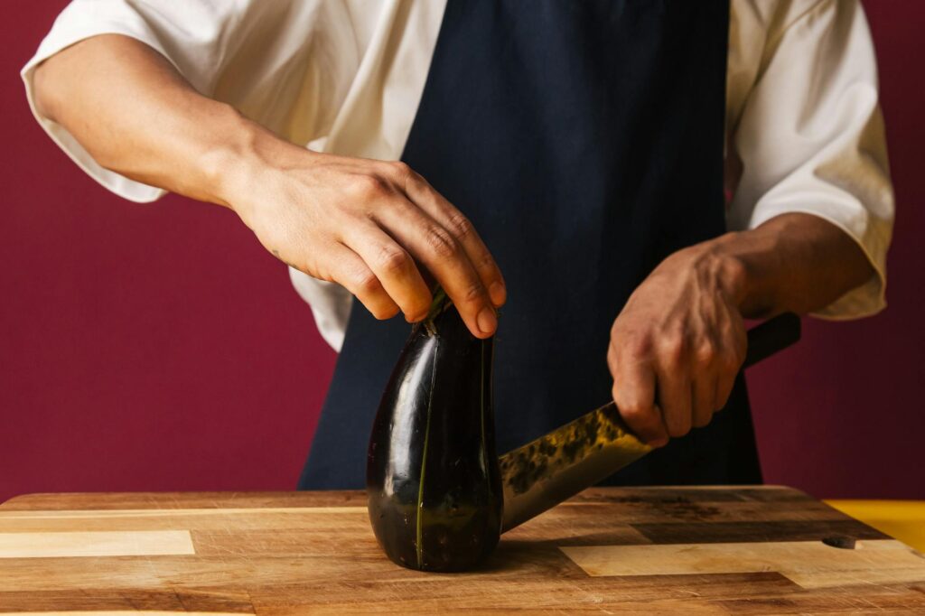 Close-up of a person's hands using a chef's knife to chop red bell peppers on a wooden cutting board.