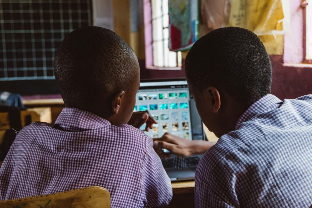 Several students gathered around a desk, pointing at a tablet and discussing a project in a sunlit classroom.