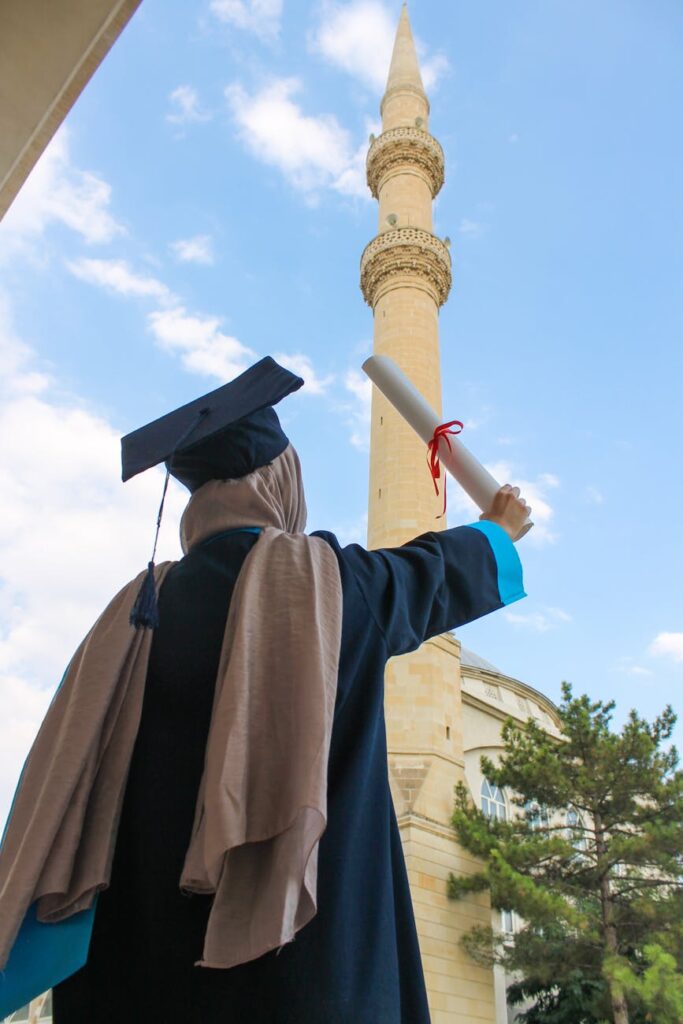 A smiling graduate student in a cap and gown proudly holding up their diploma scroll on a sunny day.