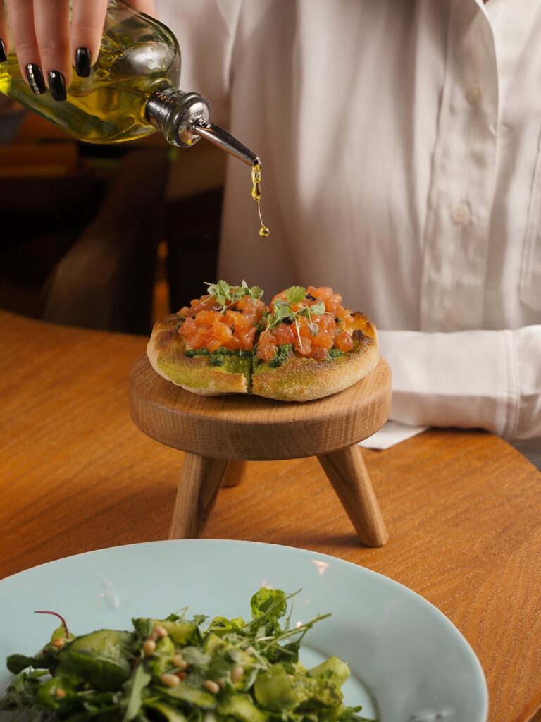 A close-up of a person's hands pouring golden extra virgin olive oil from a clear bottle onto a colorful garden salad.