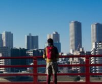 A traveler in a casual jacket looks down at a paper map while standing on a city street.
