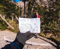 A traveler in hiking gear consults a foldable paper map while standing on a forest trail.