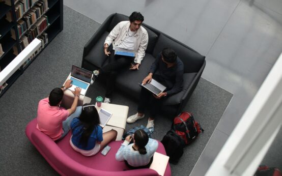 A diverse group of university students working together around a table in a sunlit library, engaged in discussion.