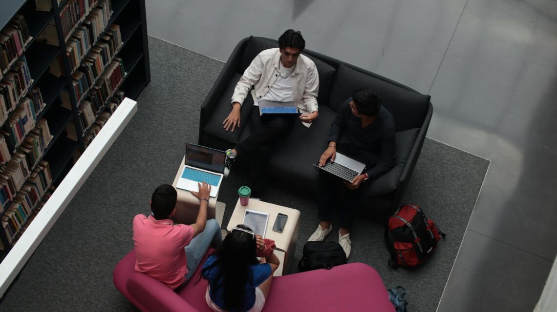 A diverse group of university students sitting at a table, focused on their work on various laptops.