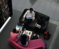 A diverse group of university students sitting at a table, focused on their work on various laptops.