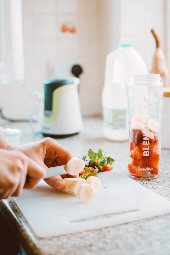A smiling person holds a glass of a vibrant green smoothie, with fresh spinach and fruits visible in the background kitchen.