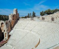 The sun-bleached stone tiers of an empty ancient Greek amphitheater, overlooking a serene landscape.