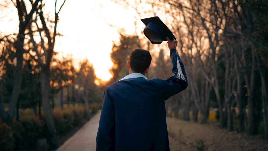 A joyful graduate in a cap and gown smiles as they toss their graduation cap into the blue sky.