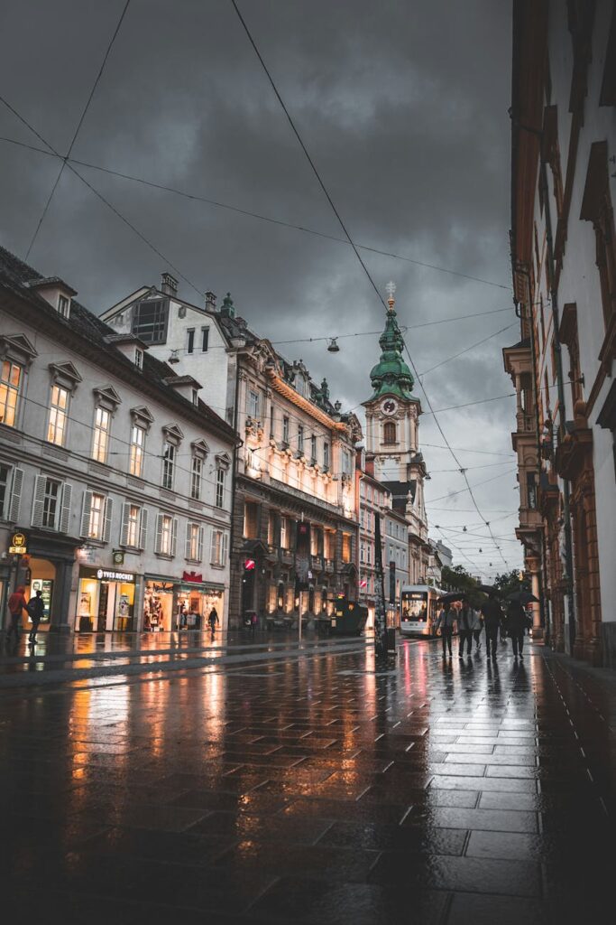 A narrow, wet cobblestone street in an old European town, illuminated by the warm glow of streetlamps as evening sets in.