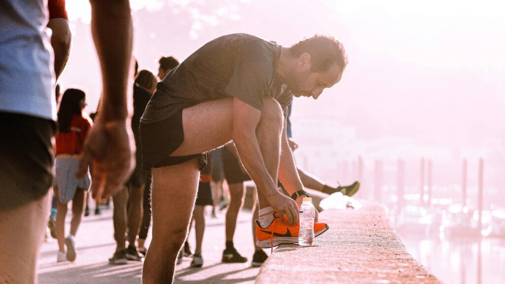An athlete in sportswear crouches to tie their shoelaces, with a blue sports drink bottle sitting on the track beside them.
