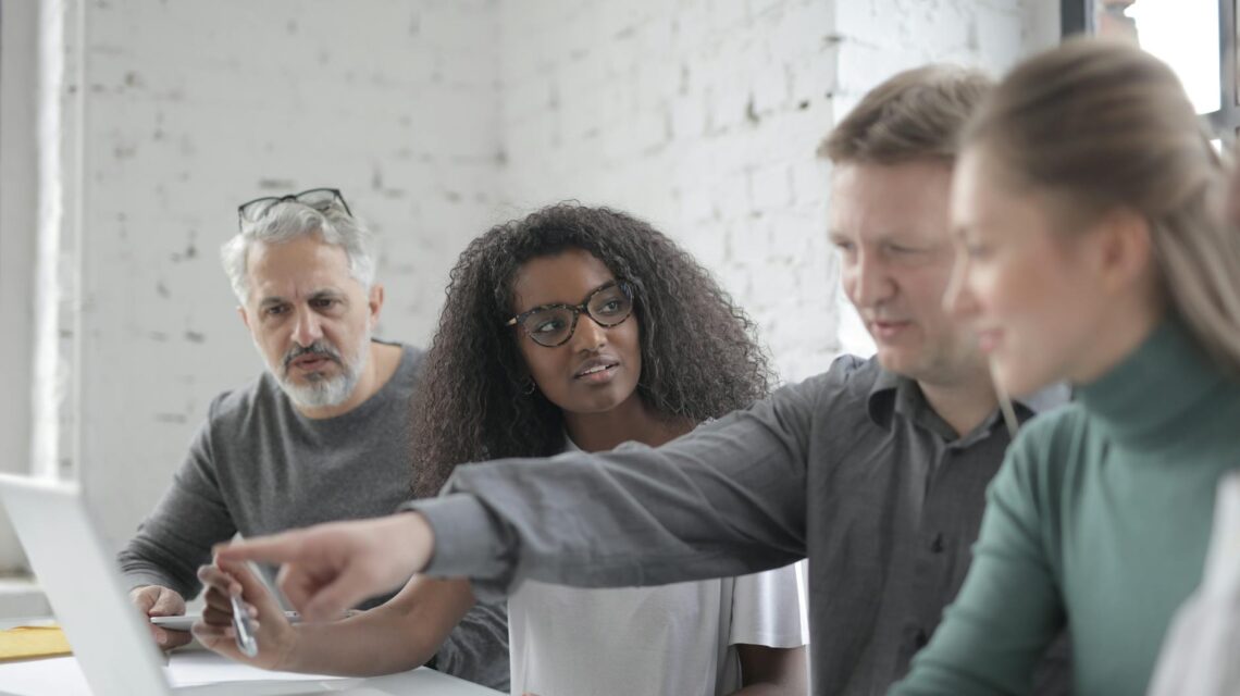 A diverse team working together around a table, pointing at a laptop screen and discussing a project.