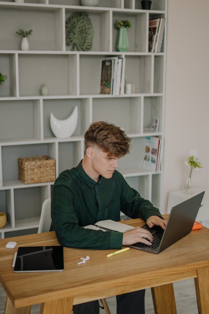 A focused student wearing headphones types on a laptop at a desk in a cozy dorm room.