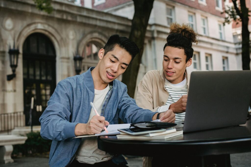 A group of four diverse students work together at a wooden table in a sunlit library, pointing at a laptop screen.