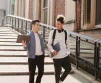 A diverse group of college students laugh and work together at a picnic table on a sunny campus green.