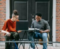 A diverse group of university students sit at a library table, focused on their laptops and books.