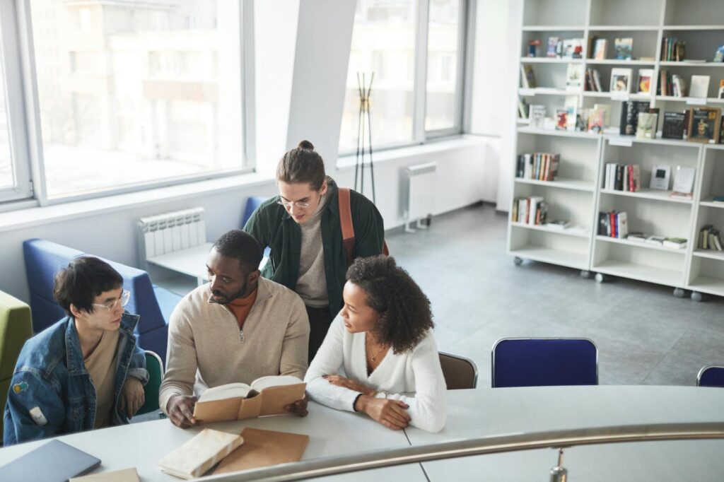 A diverse group of four smiling university students sitting on the grass and working together on a laptop.