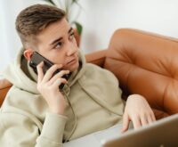 A young student sits alone in a dimly lit dorm room, the glow of their laptop screen illuminating their face.