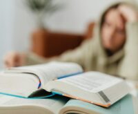 A focused student with headphones on, writing in a notebook in a sunlit library.